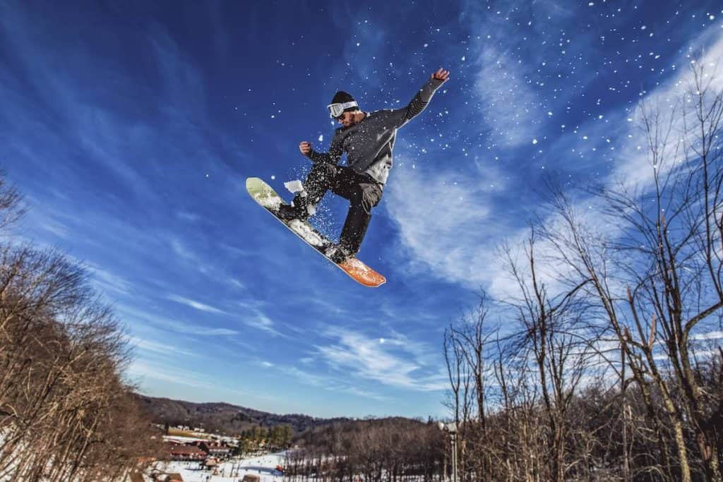Male snowboarder caught in mid-air on Appalachian Ski Mountain. Ski lodge in background at the bottom of frame. Bright blue sky behind snowborder wearing black outfit.