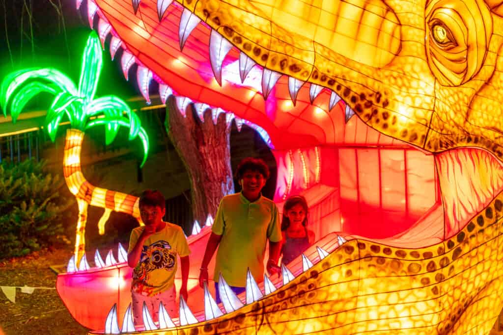 Three children in the mouth of a large illuminated dinosaur lantern at LuminoCity Festival at Pullen Park, Raleigh, NC